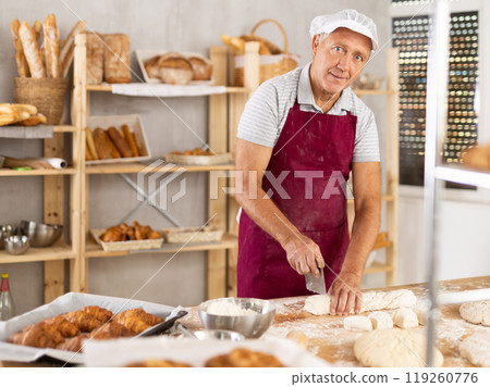 Senior man works in bakery as baker, cuts dough into portions, forms pieces of dough to create buns. Senior man works in bakery as baker, cuts dough into portions, forms pieces of dough to create buns. 119260776