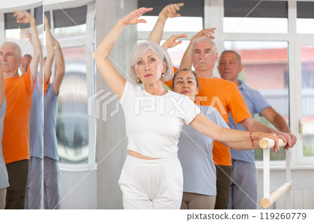 Ordinary elderly people stand in third position near ballet barre during group training in dance studio 119260779