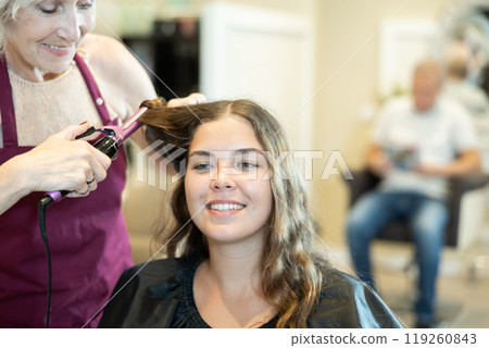 In beauty salon, elderly woman makes styling curls for young girl client. 119260843