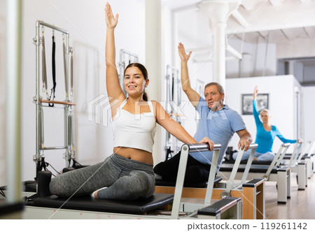 Young woman in sportswear sitting and doing Pilates exercises for hands on reformer bed equipment in fitness center 119261142