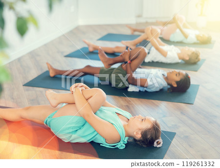 Children doing yoga with group in studio, lying on mat with knee pressed to chest with hands, performing hamstring and low back stretching Ardha Pawanmuktasana Children doing yoga with group in studio, lying on mat with knee pressed to chest with hands, performing hamstring and low back stretching Ardha Pawanmuktasana 119261332