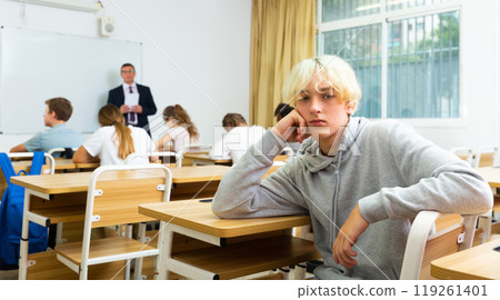 Upset teenage schoolboy sitting in classroom during lesson Upset teenage schoolboy sitting in classroom during lesson 119261401