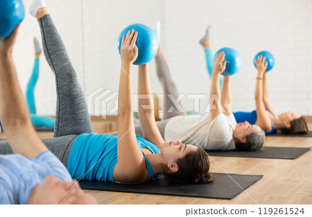 Group of young mature old people in sportswear exercising Pilates with balls in hands while lying on mats in rehabilitation center 119261524