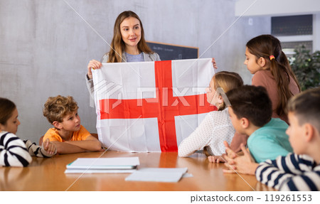 Smiling young female teacher conducting lesson for preteens, showing state flag of England in audience of middle school 119261553