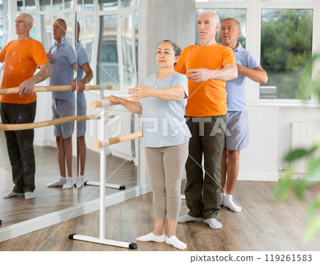 Happy mature woman in sportswear, participating first position of ballet stance at barre with group people standing in row in choreography studio Happy mature woman in sportswear, participating first position of ballet stance at barre with group people standing in row in choreography studio 119261583