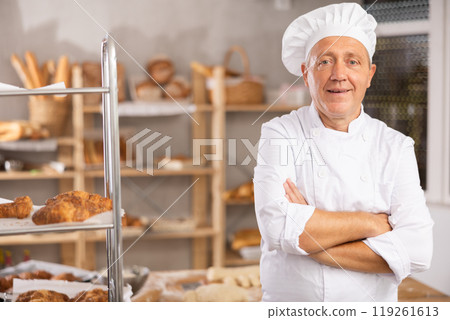 Mature man baker in white coat overall and cap stands in kitchen of bakery Mature man baker in white coat overall and cap stands in kitchen of bakery 119261613