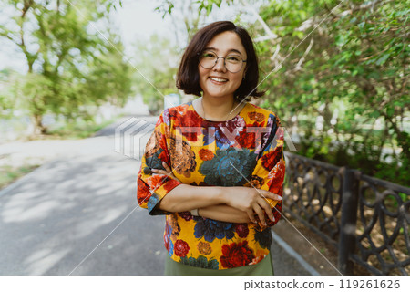 Joyful young Asian woman with crossed arms enjoying a sunny day in the park. A smiling woman with crossed arms stands in a lush park and radiates happiness Joyful young Asian woman with crossed arms enjoying a sunny day in the park. A smiling woman with crossed arms stands in a lush park and radiates happiness 119261626