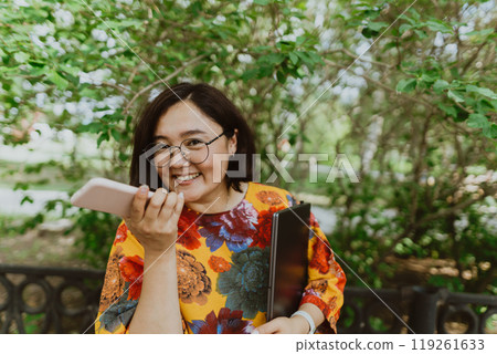 Joyful woman with smart watch and laptop talking on the phone on a sunny day in the park. Young businessman with digital devices staying connected in city green area Joyful woman with smart watch and laptop talking on the phone on a sunny day in the park. Young businessman with digital devices staying connected in city green area 119261633