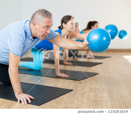 People of different ages stretching leg and arm, holding mini ball in their hands during Pilates workout in studio 119261634