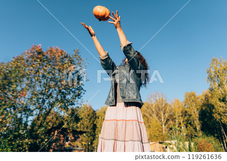 Joyful woman tossing a pumpkin on a sunny autumn day in the countryside. Middle-aged woman celebrating the harvest season in her garden with a pumpkin 119261636