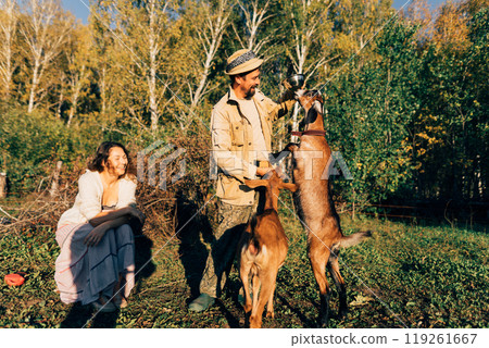 A man farmer with a beard and a hat on a small farm feeds Nubian goats on a sunny, peaceful day. Scene showcases the connection between the farmer and her animals, emphasizing eco-friendly farming 119261667
