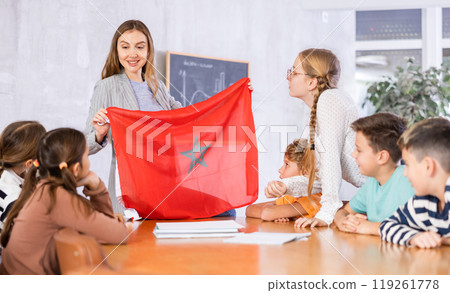 Smiling young female teacher conducting lesson for preteens, showing state flag of Morocco in audience of middle school Smiling young female teacher conducting lesson for preteens, showing state flag of Morocco in audience of middle school 119261778
