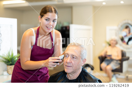 Young female hairdresser cutting hair of elderly client with clipper in salon 119262419