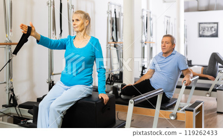 Mature woman in sportswear sitting and doing Pilates exercises for hands on reformer bed equipment in fitness center 119262433