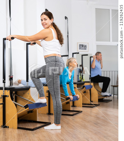 Smiling young European woman working on Pilates wanda chair machine with standing exercises during wellness training Smiling young European woman working on Pilates wanda chair machine with standing exercises during wellness training 119262435