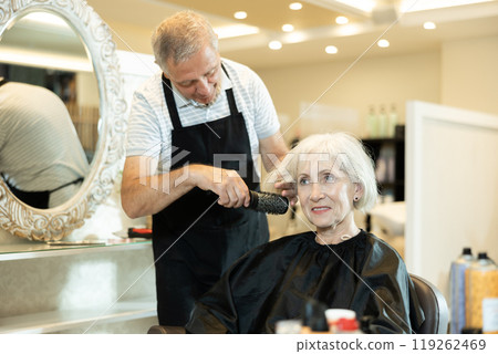 Elderly male hairdresser performs hair styling and creates hairstyle with curling iron and comb 119262469