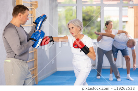 Senior lady and male couch are boxing in gym, man helps student to work out force of blow. Senior lady and male couch are boxing in gym, man helps student to work out force of blow. 119262538