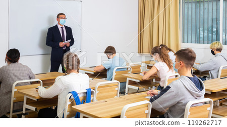 Teacher in protective mask with notebook is giving interesting lecture for students in classroom Teacher in protective mask with notebook is giving interesting lecture for students in classroom 119262717