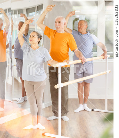 Group of positive elderly women and men engaged in classical ballet in dance studio performs exercise standing in 3rd position of ballet stance 119262832