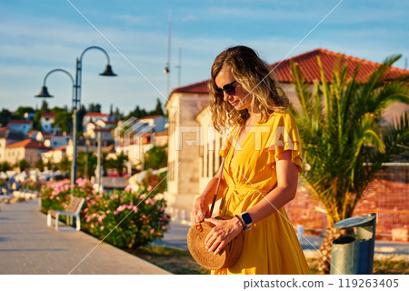 Woman is walking alone on sunny promenade with palm trees and flowers. Female tourist strolls along marina at sunset in coastal town 119263405