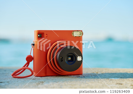 Vintage orange instant camera on rock by seaside with blurred coastal town in background. Summer travel concept. Equipment for capturing moments 119263409