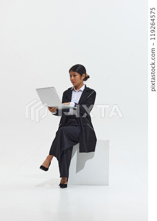 Young Asian woman in professional black outfit sits with laptop, looking focused and absorbed in her work against white studio background. 119264575