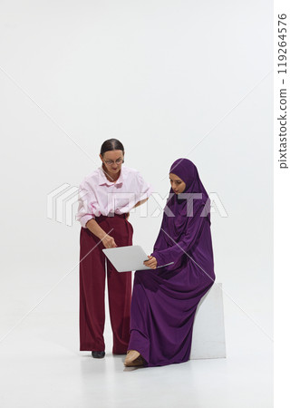 Two women collaborating on laptop. One stands, while other seated, both focused on work against white studio background. Two women collaborating on laptop. One stands, while other seated, both focused on work against white studio background. 119264576