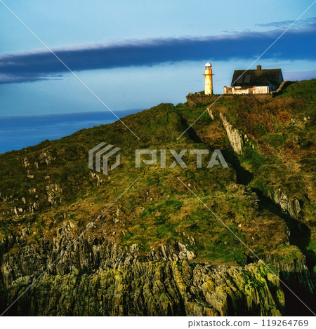 A white lighthouse stands on a rocky cliff, next to a small, white, single-story house with a grey roof. The lighthouse is tall and slender, with a white cylindrical body and a black lantern. A white lighthouse stands on a rocky cliff, next to a small, white, single-story house with a grey roof. The lighthouse is tall and slender, with a white cylindrical body and a black lantern. 119264769