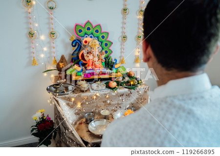 Indian man dressed in traditional wear engages in worship at home altar, adorned with decors and offerings for god Ganesh during festive celebration. Indian culture, hindu ritual and customs. 119266895