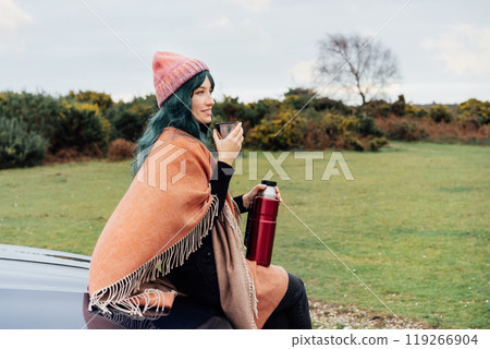 Hipster Woman sits on car's hood, wrapped in warm Shawl Wraps Poncho Cape and hat and enjoying warm drink, while overlooking rural landscape during road trip. Cold season travel, vacation concept 119266904