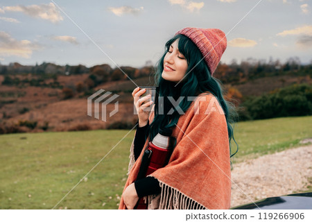 Hipster woman wrapped in Poncho with closed eyes enjoying moment with warm drink on car hood while taking in serene rural landscape during cold season road trip. Fall, winter travel, vacation concept. 119266906