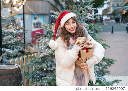 Young woman in Santa hat with with a tower of gift boxes on traditional city street Christmas market. Happy woman choosing Christmas gifts. Merry Xmas Happy New Year festive shopping, sales, discount. 119266907