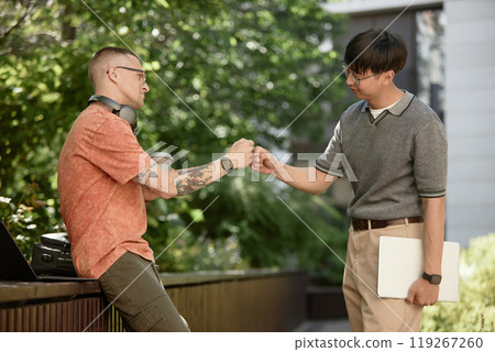 Side view of IT programmer bumping fists with partner making deal while having fun on meeting in park, copy space 119267260