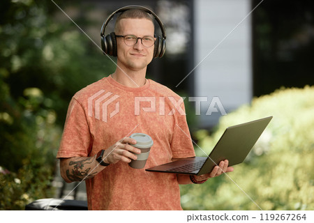 Medium portrait shot of smiling businessman in headphones holding laptop and reusable cup captured during outdoor freelance work in park, copy space Medium portrait shot of smiling businessman in headphones holding laptop and reusable cup captured during outdoor freelance work in park, copy space 119267264