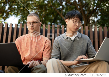 Medium shot of two male IT developers concentrated on writing code on laptops while working together sitting on bench outdoors in city park, copy space 119267296