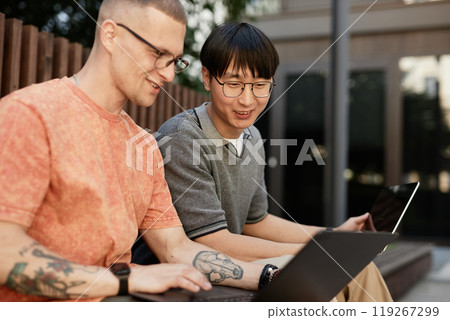 Candid shot of smiling Asian male IT programmer working on laptop collaboratively with colleague having fun while sitting on bench outside in city street, copy space Candid shot of smiling Asian male IT programmer working on laptop collaboratively with colleague having fun while sitting on bench outside in city street, copy space 119267299