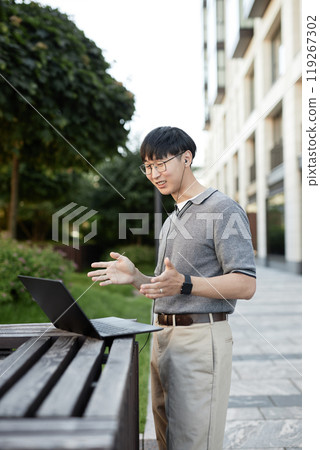 Vertical shot of smiling Asian businessman looking at laptop screen talking with colleague captured during online conference while working at stand up desk outside in city street, copy space Vertical shot of smiling Asian businessman looking at laptop screen talking with colleague captured during online conference while working at stand up desk outside in city street, copy space 119267302