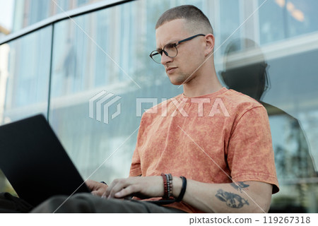 Medium shot of serious male software engineer with septum piercing and glasses typing on laptop while sitting outside leaning against glass construction in city street, copy space Medium shot of serious male software engineer with septum piercing and glasses typing on laptop while sitting outside leaning against glass construction in city street, copy space 119267318