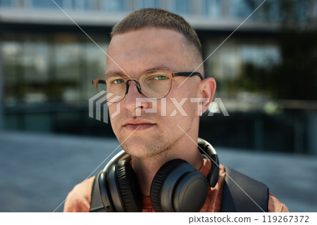 Portrait of confident adult man with headphones wearing glasses standing outside in city street while looking at camera on blurred background, copy space Portrait of confident adult man with headphones wearing glasses standing outside in city street while looking at camera on blurred background, copy space 119267372