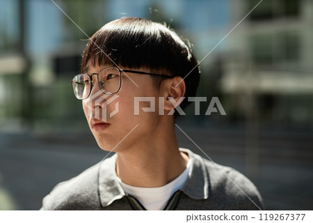 Close up of adult Asian man with glasses looking away lost in thought while standing outside in sunlight on blurred background, copy space 119267377