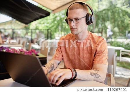 Medium shot of concentrated businessman in wireless headphones using laptop to work or study typing on keyboard while sitting at table in outdoor seating area of cafe, copy space Medium shot of concentrated businessman in wireless headphones using laptop to work or study typing on keyboard while sitting at table in outdoor seating area of cafe, copy space 119267401