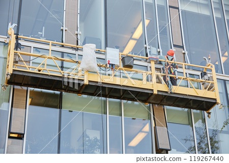 Background shot of building cradle hanging outside business building while worker installing facade elements, copy space 119267440