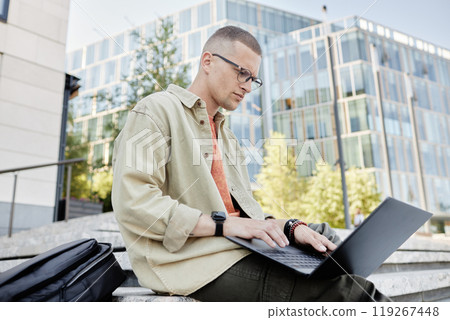 Medium shot of serious man with glasses and tiny septum using laptop for working or studying outdoors while sitting outside in city street, copy space 119267448