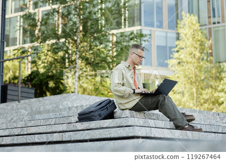 Full shot of concentrated male IT programmer in casual clothing using laptop to study or write code sitting on concrete steps with backpack lying next to man outside in city, copy space 119267464