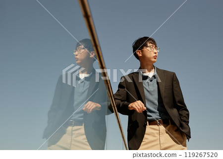 Medium shot of stylish Asian businessman wearing black jacket enjoying sunlight outside in city while standing next to reflective glass construction against blue sky, copy space Medium shot of stylish Asian businessman wearing black jacket enjoying sunlight outside in city while standing next to reflective glass construction against blue sky, copy space 119267520