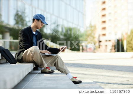 Side view of Asian male IT programmer writing code on laptop looking at computer screen while sitting on concrete steps in city street, copy space 119267560