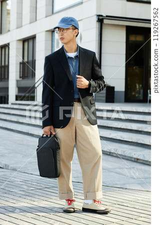 Vertical shot of fashionable Asian man wearing black jacket outfit and holding laptop bag while posing in city street captured during daily commute on foot to workplace, copy space Vertical shot of fashionable Asian man wearing black jacket outfit and holding laptop bag while posing in city street captured during daily commute on foot to workplace, copy space 119267562