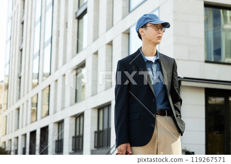 Medium shot of fashionable Asian man wearing black jacket outfit for office or business meeting keeping one hand in pocket while standing outside in city street, copy space Medium shot of fashionable Asian man wearing black jacket outfit for office or business meeting keeping one hand in pocket while standing outside in city street, copy space 119267571