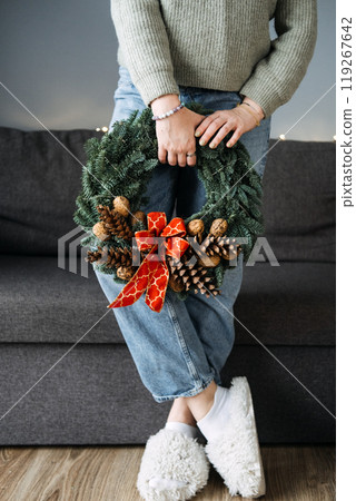 Person holding handcrafted Christmas wreath with pinecones and red bow, standing in cozy living room. DIY holiday traditions, handmade decor, and personal creativity. Person holding handcrafted Christmas wreath with pinecones and red bow, standing in cozy living room. DIY holiday traditions, handmade decor, and personal creativity. 119267642