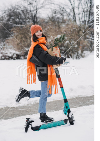 Smiling woman on e-scooter in snowy park wearing orange scarf and beanie, holding festive gift bag. Urban winter eco-commuting during holiday shopping season 119267699
